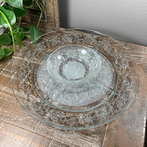 May include: Clear glass serving set with a large plate, a smaller plate, and a bowl. The plates and bowl have a floral pattern around the edges and in the center. The set is on a wooden surface.