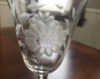May include: A clear glass stemmed wine glass with an etched floral design. The design features a large flower with a geometric center and smaller leaves.