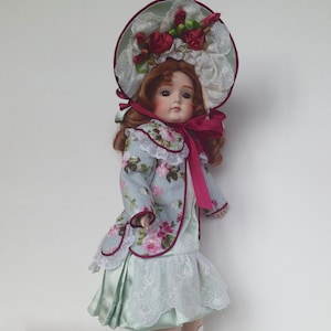May include: A porcelain doll with red hair, wearing a light blue dress with floral print and a white lace trim. The doll is wearing a pink ribbon around her neck and a white hat with red roses and lace.