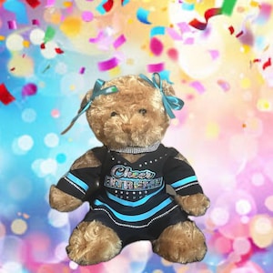 May include: A tan plush teddy bear wearing a black cheerleading uniform with bright blue stripes and the text Cheer Extreme across the chest. The uniform is accented with silver rhinestones, and the bear has two light blue ribbons on its head.