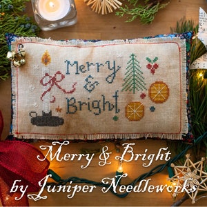 May include: A cross-stitch pillow with the text "Merry & Bright" and a design of a candle, a Christmas tree, and oranges. The pillow is on a wooden surface with Christmas lights, pine branches, and other decorations.