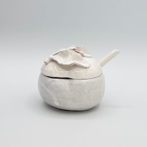 May include: A small, off-white ceramic sugar bowl with a lid featuring a floral design. The bowl has a rounded shape and a small handle. The ceramic has a crackled glaze.