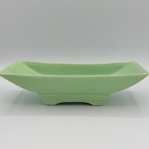 May include: A pale green ceramic dish with a rectangular form and angled sides. The dish has a speckled texture and rests on a low, stepped base. The design is minimalist and modern.