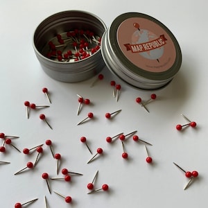 May include: A silver tin with a red and white label that says "MAP REPUBLIC" and a website address. The tin is filled with red push pins. Scattered around the tin are more red push pins on a white surface.