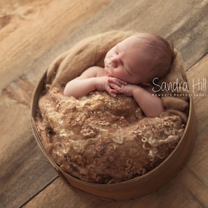 May include: A newborn baby sleeping peacefully in a brown woven basket, wrapped in a fluffy, cream-colored blanket. The baby's eyes are closed and their hands are clasped together. The basket is sitting on a wooden surface.