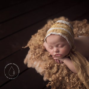 May include: A newborn baby wearing a white and yellow crocheted bonnet sleeps on a brown fluffy blanket. The baby is wearing a matching yellow and white crocheted shawl.