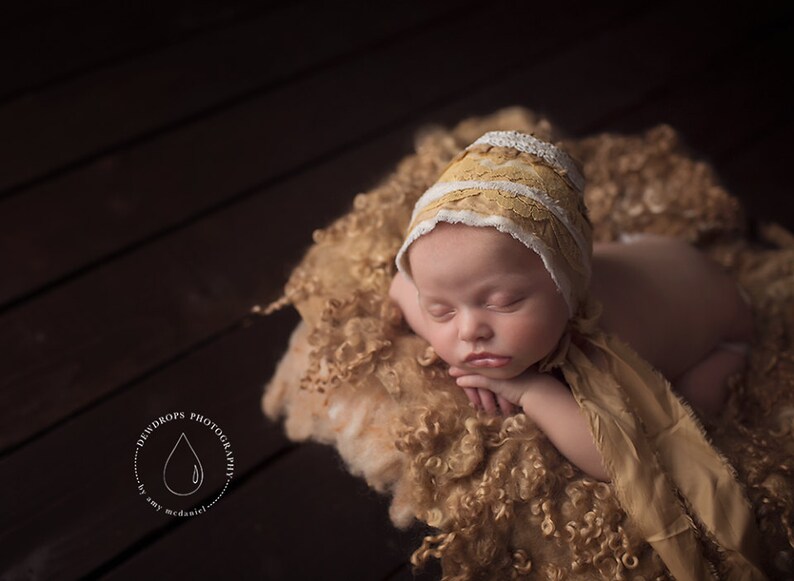 May include: A newborn baby wearing a white and yellow crocheted bonnet sleeps on a brown fluffy blanket. The baby is wearing a matching yellow and white crocheted shawl.