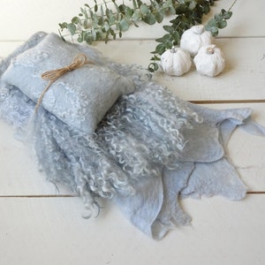 May include: A light blue, fluffy, and textured baby blanket with a smaller, light blue, patterned pillow tied with twine. The blanket is folded and arranged on a white wooden surface.