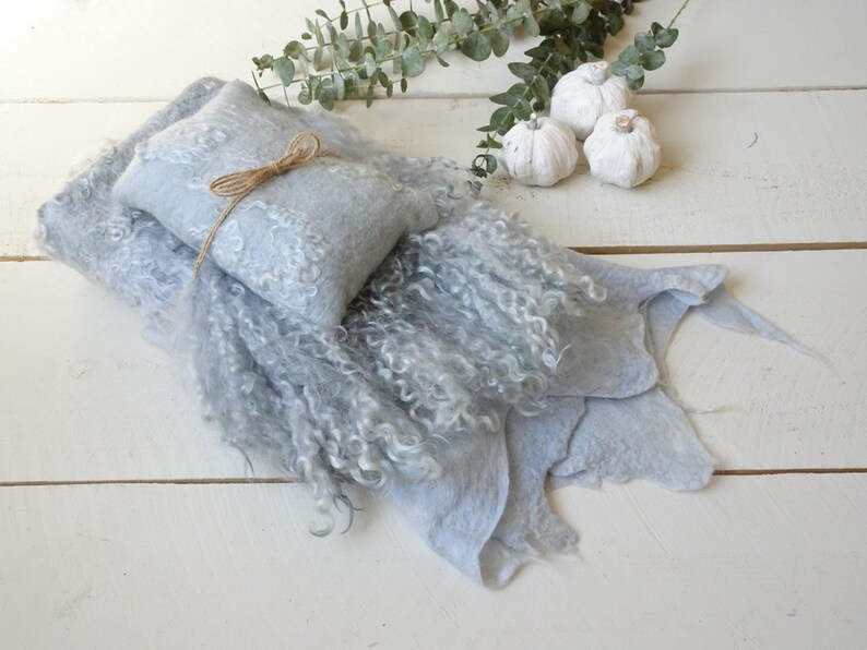 May include: A light blue, fluffy, and textured baby blanket with a smaller, light blue, patterned pillow tied with twine. The blanket is folded and arranged on a white wooden surface.