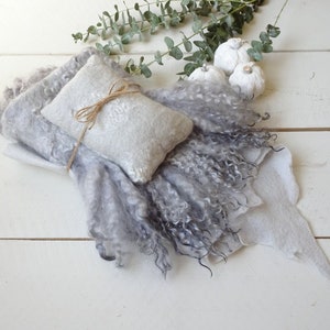 May include: A gray and white fluffy blanket with a smaller gray and white pillow tied with twine. The blanket and pillow are on a white wooden surface with white pumpkins in the background.