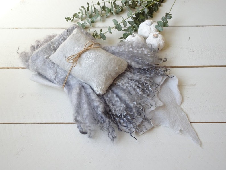 May include: A gray and white fluffy blanket with a smaller gray and white pillow tied with twine. The blanket and pillow are on a white wooden surface with white pumpkins in the background.