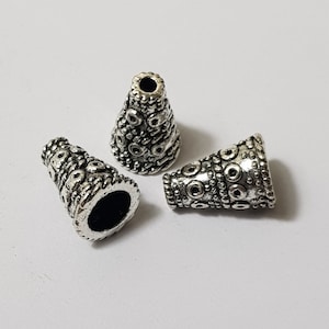 May include: Three silver metal cone-shaped bead caps with an intricate embossed design. The caps have a circular opening at the top and a smooth, flat bottom.