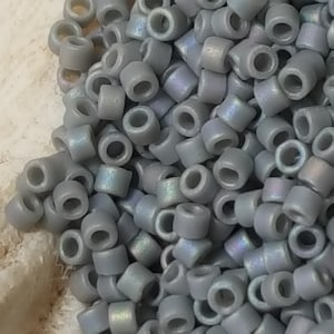 Puede incluir: Un montón de pequeñas cuentas de vidrio grises e iridiscentes. Las cuentas son redondas y tienen un agujero en el centro.