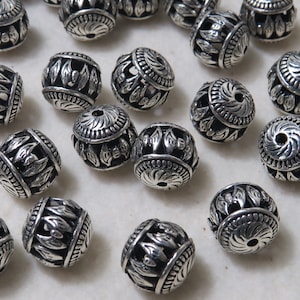 Könnte beinhalten: Eine Sammlung silberfarbener Metallperlen mit filigranen Blumendesigns. Die Perlen sind rund und haben eine glatte, polierte Oberfläche.