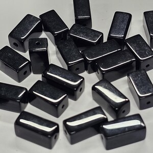 May include: A collection of rectangular black beads with a smooth, glossy finish. Each bead features a single drilled hole, reflecting light on its surface. The beads are scattered in a group, showcasing their uniform shape and dark, reflective colour.