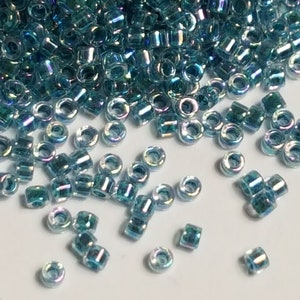 Può includere: Un mucchio di piccole perline di vetro blu iridescente. Le perline sono rotonde e hanno un foro al centro. Sono sparse su una superficie bianca.