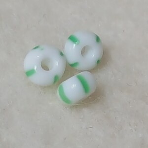 Pu&ograve; includere: Tre perline di vetro bianco con strisce verdi. Le perline sono rotonde e hanno un foro al centro.