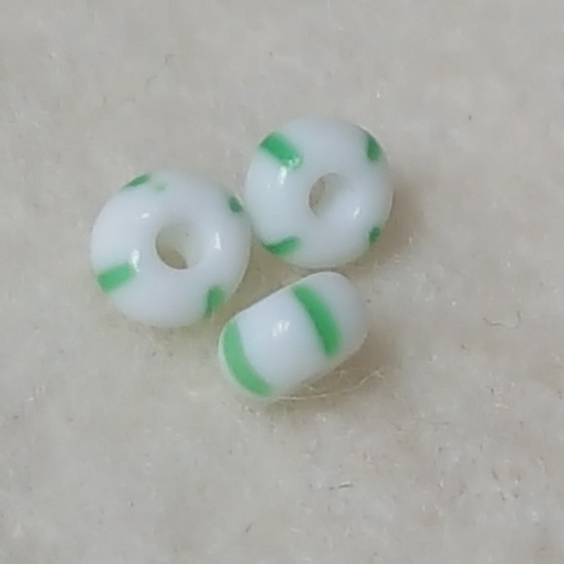 Pu&ograve; includere: Tre perline di vetro bianco con strisce verdi. Le perline sono rotonde e hanno un foro al centro.