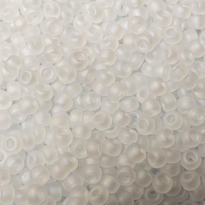 May include: A close-up view of a pile of small, white, opaque seed beads. The beads have a slightly iridescent finish.