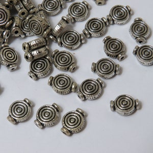 May include: A collection of silver metal beads with a circular design. The beads have a concentric circle pattern.