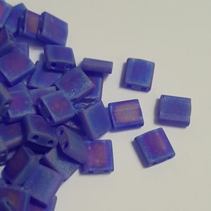 May include: A pile of small, square, iridescent blue glass beads with two holes each. The beads have a slightly textured surface and are scattered on a white background.