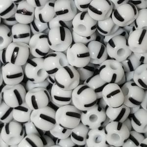 May include: A close-up image of white round beads with black stripes. The beads are small and have a hole in the centre.