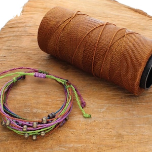 May include: A colourful multi-strand bracelet with green, purple, blue, and orange cords, featuring small metallic beads. A spool of brown cord is in the background. The bracelet is on a wooden surface.