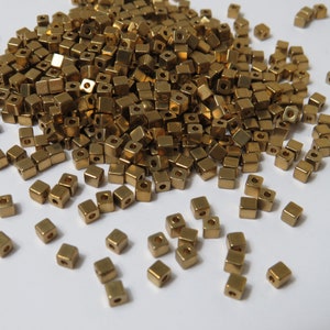 May include: A large pile of gold-coloured metal square beads. The beads are scattered on a white surface.