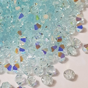 May include: A pile of light blue crystal beads with a faceted design. The beads are scattered on a white surface.