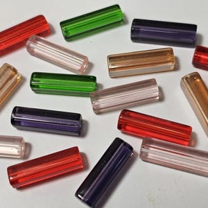 May include: A collection of colourful, rectangular glass beads in shades of red, green, purple, pink, and gold. Each bead has a ribbed texture and a translucent appearance, ideal for jewellery making or craft projects.
