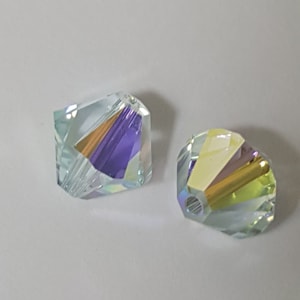 May include: Two iridescent glass beads with a faceted design. The beads are light blue with a purple and gold iridescent sheen.
