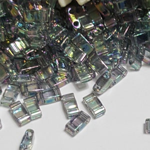 Pode incluir: Um monte de contas de vidro iridescentes prateadas e pretas. As contas são retangulares e têm uma superfície plana e lisa. São perfeitas para fazer joias ou outros projetos de artesanato.