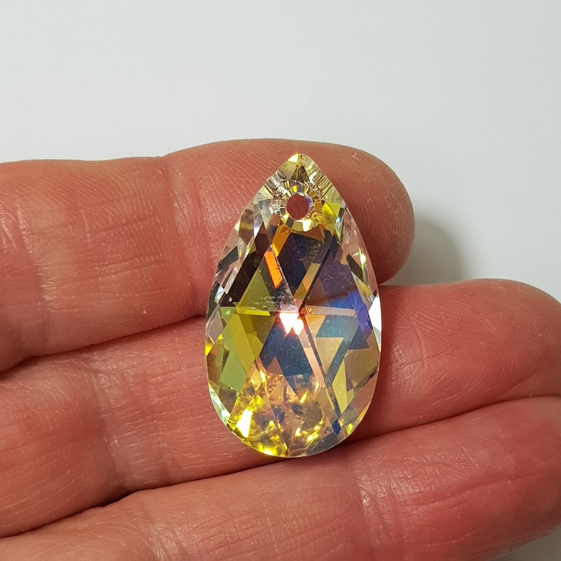 May include: A teardrop-shaped crystal pendant with a faceted design and a hole at the top for hanging. The crystal is iridescent and has a yellow hue.