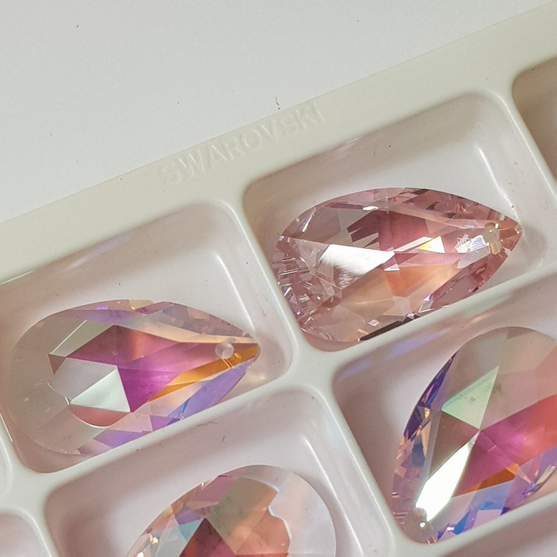 May include: A close-up of a clear plastic container with several pink iridescent crystal teardrop shapes. The container has the word "SWAROVSKI" printed on the top.
