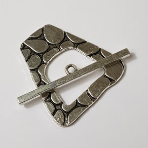 May include: Silver-toned metal buckle with a textured, pebble-like design. The buckle has a rectangular frame with a rounded edge and a straight bar that crosses through the frame.