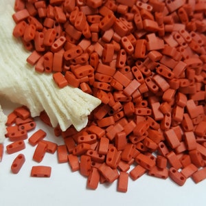 Può includere: Un grande mucchio di piccole perline rettangolari di colore rosso-arancio. Le perline hanno due fori ciascuna. Le perline sono sparse su una superficie bianca.