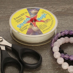 Könnte beinhalten: Eine Spule weißer elastischer Stretch Magic Schnur, 8 mm im Durchmesser, mit einem gelben Etikett mit der Aufschrift "Stretch Magic" und "25 Meter/82 Fuß". Das Etikett enthält auch den Text "Bead & Jewelry Cord, Perle & Cordon de Bijouterie, strong & stretchy/fort & elastique".