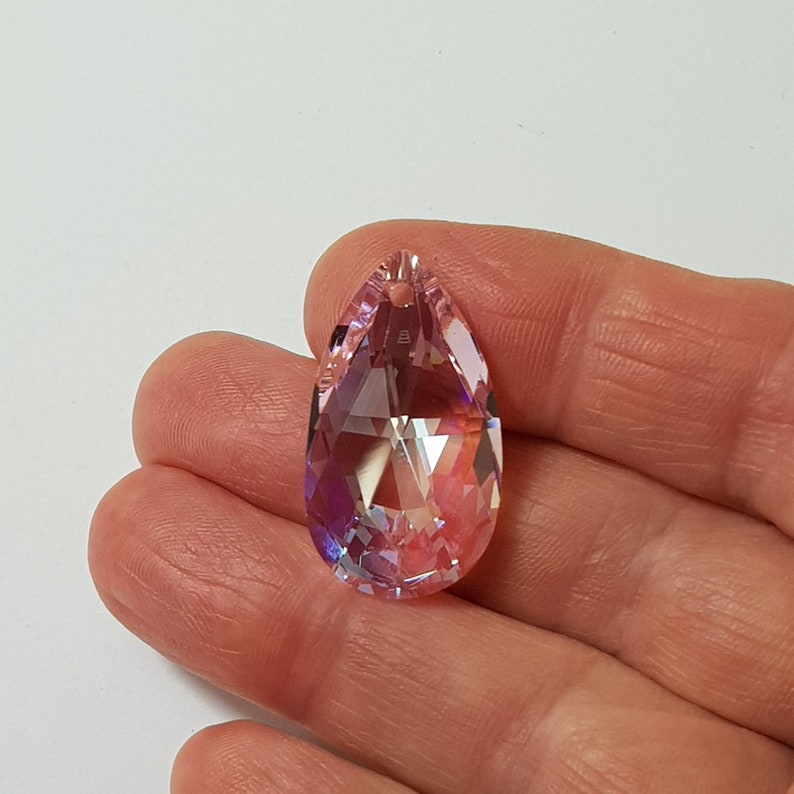 May include: A teardrop-shaped crystal pendant with a pink iridescent finish. The pendant has a faceted design and a hole at the top for hanging.
