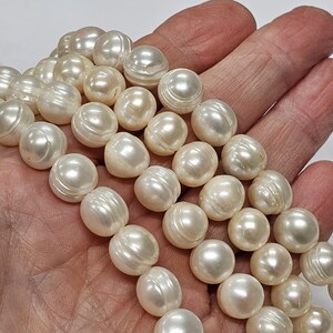 Pode incluir: Um colar de pérolas redondas de cor branco-sujo com um brilho lustroso e texturas superficiais naturais visíveis. As pérolas estão dispostas em várias filas, exibindo o seu acabamento liso e refletor, bem como o seu tamanho consistente.
