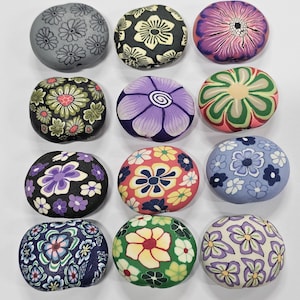 May include: A selection of twelve round beads, each decorated with a floral pattern. The beads display intricate flower designs in shades of purple, pink, green, yellow, and blue, set against black, grey, and white backgrounds.