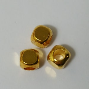 May include: Three gold-coloured metal beads with a square shape and a hole in the centre. The beads are on a white surface.