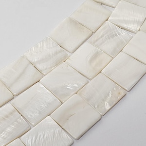 May include: Close-up of a strand of square, off-white mother-of-pearl beads. Each bead has a slightly iridescent surface, reflecting light. The beads are arranged in a diagonal pattern, showcasing their smooth, polished texture. Ideal for jewelry making.
