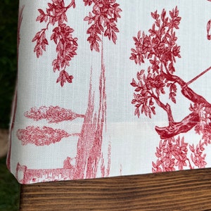 Pu&ograve; includere: Primo piano di un tessuto bianco con un motivo toile rosso che presenta alberi e un paesaggio. Il tessuto &egrave; piegato su una superficie di legno.
