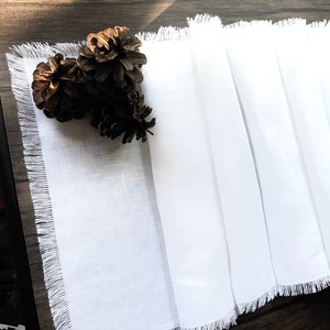 May include: White linen table runner with frayed edges. The runner is folded in half and placed on a wooden table. Three pine cones are placed on top of the runner.