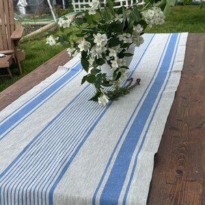 May include: A linen table runner with blue stripes on a gray background. A white vase with white flowers and green leaves sits on the runner, which is placed on a dark wooden table. The scene is outdoors.