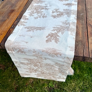 May include: A rectangular table runner with a light blue background and a brown toile pattern featuring trees and landscapes. The runner is draped over a wooden table, with green grass visible in the background.