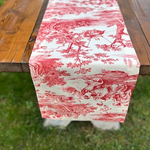 Pu&ograve; includere: Un runner da tavolo in toile rosso e bianco con un design a tema caccia. Il runner &egrave; su un tavolo di legno.