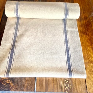 May include: A white linen table runner with two blue stripes down the sides. The runner is rolled up on a wooden table.