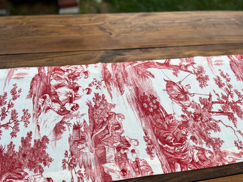 Pu&ograve; includere: Un runner da tavolo rosso e bianco con un motivo toile che rappresenta una scena pastorale con alberi, persone e animali.