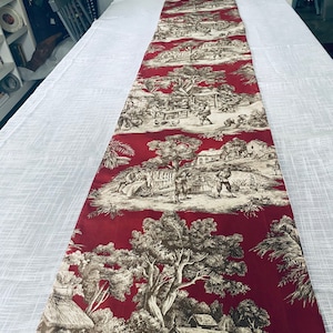 May include: A red table runner with a white background featuring a repeating pattern of black and white stylized trees, houses, and figures. The pattern is reminiscent of a toile design.